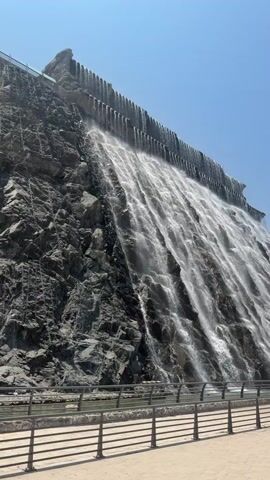 A large, man-made waterfall cascades down a steep, dark grey rocky slope under a clear blue sky. Water flows in numerous streams over the textured rock face, originating from a horizontal structure at the top. At the base of the waterfall, a narrow body of water is visible. A light-colored concrete pathway or promenade runs along the edge of the water, separated by a black metal railing. The camera slowly pans horizontally to the right, maintaining a view of the waterfall and its surroundings. No people or animals are visible.