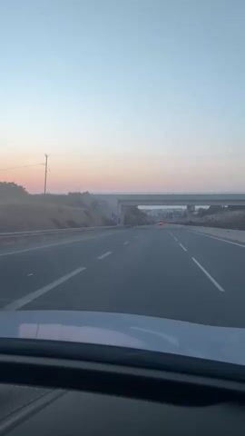 This is a POV video from a car driving on a highway. The road has multiple lanes and is bordered by a median with a guardrail and dry, grassy hills. An overpass with graffiti is visible in the distance. The sky is clear with a gradient of light blue to pale pink, indicating dawn or dusk. Two cars with red taillights are visible on the road ahead. The interior of the car, including the dashboard, is visible in the foreground.