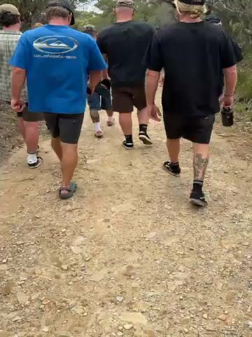 The video shows a group of seven people walking away from the camera on a dirt path. They are dressed casually in shorts and t-shirts or button-down shirts. Some are wearing sneakers, and at least one person is carrying a water bottle. The path appears to be in a natural, wooded area.