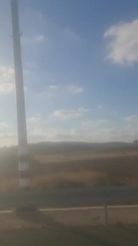 The video is an unedited, vertical, outdoor shot of a rural landscape. The top two-thirds of the frame are filled with a bright blue sky dotted with white, wispy clouds. The bottom third of the frame depicts a hazy, brown-toned landscape, likely fields, with a line of trees or dense shrubbery visible in the distance. The sun appears to be low on the horizon, casting a soft light and creating a hazy effect. A faint, dark horizontal line, possibly a road or a fence, runs across the lower portion of the frame. There are no discernible people, specific objects, or actions occurring within the video. The scene conveys a sense of open space and natural scenery.