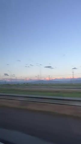 The video shows a landscape with mountains in the background. The peaks of the mountains are covered in snow and are illuminated by the warm light of the setting or rising sun. A highway runs in the foreground, with a median and some grass and a fence. Streetlights line the road. Power lines are visible stretching across the horizon. The sky is a clear blue with a few small clouds. The video is shaky, suggesting it was taken from a moving vehicle. There are voices heard in the background.