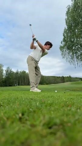 A man is playing golf on a green golf course on a cloudy day. He is wearing a light-colored sweater, khaki pants, and light-colored shoes. He is standing with his back to the camera, holding a golf club, and watching the golf ball he just hit. There is a tree to the right of the frame, and other trees and green hills in the background. The camera is low to the ground, focusing on the grass.