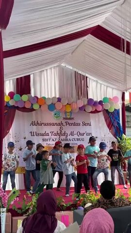 A group of young boys are performing a synchronized dance on a raised stage under a tent. The stage is decorated with red and white draping and colorful balloons. A banner behind the performers reads "Akhirussanah, Pentas Seni & Wisuda Tahfidz Al-Qur'an SDIT AZ-Zahra Curug Tahun Ajaran 2022-2023." The boys are wearing various casual outfits, including t-shirts, jeans, and one boy in a green outfit. Some wear baseball caps. The audience members are visible in the foreground, some wearing headscarves. The event appears to be a school performance or graduation ceremony.