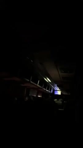 The video shows the interior of a dimly lit vehicle, likely a bus or train, moving through a dark environment. The aisle is visible with seats on either side, occupied by people whose faces are not discernible due to the low light. Overhead compartments and lights are present, with a small screen visible in the distance, displaying some content. The lighting shifts slightly throughout the clip, revealing glimpses of the occupants and the interior structure. The overall impression is one of travel in low visibility conditions.