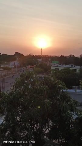 The video shows a sunset in a suburban neighborhood. The sun is visible in the sky, shining brightly and casting a warm orange glow over the surroundings. Below the sun, a cell phone tower stands tall, slightly obscuring the horizon. The foreground features lush green trees with dense foliage, partially blocking the view of houses and buildings in the neighborhood. A road is visible, with a vehicle passing. The sky is clear, with faint hues of pink and orange blending into the blue.  The camera is steady, capturing the serene atmosphere of the setting. The text "Infinix NOTE 40Pro" is visible at the bottom left corner of the frame, indicating the device used to record the video.