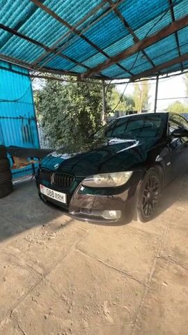A black BMW coupe is parked under a blue corrugated metal roof. The car has tinted windows and black rims. The ground is concrete and dusty. There is a fence in the background with trees and laundry hanging on a line.