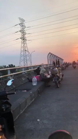 The video shows a street scene at sunset. A high-voltage electrical tower is visible in the background, with power lines stretching across the frame. A bridge or overpass is in the foreground, with a railing and a sound barrier. Several motorcycles are parked along the side of the road. A person is sitting on the edge of the bridge, appearing to be engaged in an activity, possibly eating or working on something. Other individuals are standing near the railing, observing the surroundings. The sky is filled with the warm hues of a sunset, casting a soft light over the scene. The overall atmosphere appears to be one of a peaceful evening, with people gathering or pausing on their way.