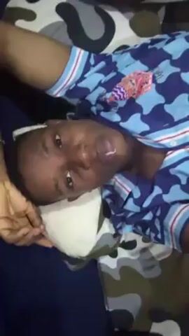 A person lies on a bed, looking at the camera. The person is wearing a blue and white patterned shirt. The bed has a gray, brown, black, and white camouflage pattern. The person's left arm is resting on a white pillow. The bed also has a dark blue part visible. The person is centered in the frame.