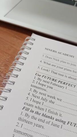 The video shows a notebook open to a page titled "NINERS ACADEMY" with exercises on the future perfect tense. A blue pen lies on the notebook. A laptop is in the background, displaying a login screen with a code. The desk is wooden. Other papers are scattered around the notebook. The camera zooms out to show the entire setup.