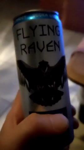 The video shows a person holding a can of "Flying Raven" drink. The can has a silver background, black lettering, and a black silhouette of a raven. The top of the can is blue. The person then opens the can, and a small amount of vapor is released. The camera then focuses on the blue top of the can, which appears to be glowing.