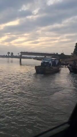 The video shows a flat-bottomed boat carrying two pickup trucks across a body of water. Several people can be seen on the boat with the vehicles and on the shore next to other boats. In the background is a bridge with its central span missing. The sky is overcast.