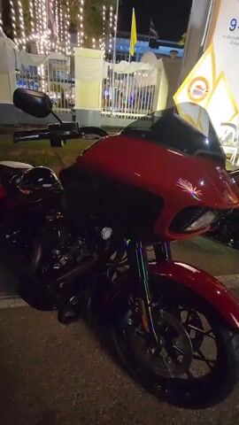 A dark-colored motorcycle with a red front fairing and a black windshield is parked on a paved surface at night. The motorcycle is positioned at an angle, with its front wheel facing the right side of the frame. Decorative lights, appearing as strings of small bulbs, are visible in the background, illuminating white tents or structures. A yellow flag and a partially visible sign are also present in the background. The lighting conditions suggest it is nighttime, with artificial illumination from the decorative lights and possibly other sources. No people are visible in the frame.