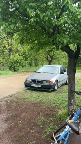 The video shows a silver BMW E36 parked near a tree. The car has a matte black hood, yellow tinted headlights, and a Bulgarian license plate that reads "CO 2710 BT". The car is parked on a grassy area next to a dirt road. A bicycle is parked near the tree. The background features lush green trees and bushes, indicating a natural, possibly rural environment. The lighting suggests an overcast or partly cloudy day.