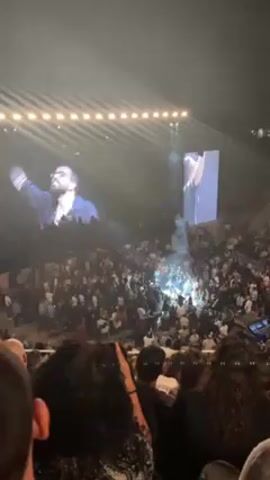 A large crowd of people is gathered in an amphitheater to watch a concert. On a large screen, a man with a beard and glasses is seen singing and drinking from a water bottle. Another, more slender screen shows him singing at the right. Spotlights shine down on the crowd from above. Many audience members hold up their phones to record the performance. The person recording the video is sitting in the crowd, with the heads and shoulders of people in front of them visible.