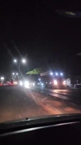 The video shows a nighttime view from inside a vehicle moving along a wet road. Streetlights illuminate the road and the surrounding area. On the right side of the road, a large truck with illuminated lights is passing by. Trees and some buildings are visible in the background. The road appears to have some puddles or wet patches reflecting the lights. The overall scene suggests a journey at night, possibly in or near a town or city.