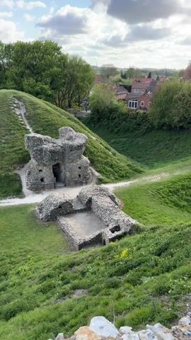The video presents a panoramic view of an ancient stone ruin and its surrounding landscape in King's Lynn and West Norfolk, United Kingdom, under a sky with both blue patches and fluffy white and grey clouds.

From 0:00 to 0:02, the camera captures the crumbling remains of a stone structure, possibly a castle or fort, situated within a grassy, undulating terrain. This main ruin features an arched opening and is surrounded by a path, with a smaller, lower stone foundation visible in front of it. Modern houses are nestled among trees in the background.

From 0:02 to 0:10, the camera pans across the expansive landscape from an elevated vantage point. The foreground is dominated by a textured stone wall, covered with moss and lichen. Beyond this, the scene unfolds into vast green fields and meadows, dotted with trees and distant woodlands under the bright, partly cloudy sky. A small wooden bridge structure is also visible in the mid-ground of the landscape. The setting appears serene and pastoral. There are no people or discernible text visible in the video.