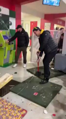 The video shows a man using a large red mallet to strike a strength test game, with the camera shifting to capture the surrounding environment, which appears to be an indoor venue with red and green decor, an ATM, and other people passing by. The man in the video is wearing a black jacket with yellow accents, and a baseball cap. The strength test game consists of a small platform and a lever. Another man, wearing a black jacket and white sneakers, is standing near the ATM, observing. The floor is littered with debris from previous attempts at the game.  A TV screen is visible in the background, showing a colorful video game.