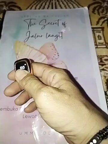 The video shows a close-up of a hand holding a small, square electronic device with a screen displaying "60" and then "61". The hand is also wearing a black beaded bracelet with a yin-yang symbol. In the background, there is a book with the title "The Secret of Jalur Langit" visible, featuring a watercolor-style illustration of butterflies and what appears to be wings. The book cover has text that seems to be in Indonesian. The overall environment appears to be indoors, with a patterned surface visible beneath the book, possibly a mat or cloth.