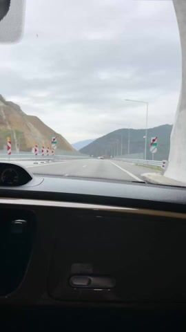 The view is from the inside of a gold-colored car dashboard as it drives along a multi-lane highway.  Mountains flank either side of the highway, receding into the distance under a cloudy sky. Guardrails and highway lights line the road. There's some traffic in the distance. The car is moving at a steady speed, with the Turbo gauge visible. There's a small yellow toy figure on the dashboard near the windshield.  Background music plays.