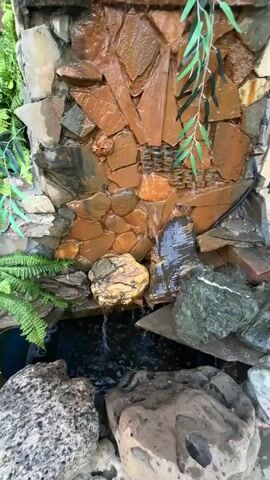 A close-up shot shows a rock garden water feature where water cascades down a wall constructed of various stones. The stones are in shades of brown, gray, and tan, with some appearing wet from the flowing water. Green ferns and a dangling branch with green leaves are visible on the sides. The water collects in a dark pool at the bottom, surrounded by large rocks. The lighting suggests an outdoor setting, likely during the day. The overall scene depicts a tranquil and naturalistic element in a garden environment.
