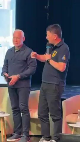 Two men stand on a stage. The man on the right is speaking into a microphone and gesturing with his right hand. He is wearing a black polo shirt with an emblem on the left breast, black pants, and a black watch on his left wrist. The man on the left is listening and holding a tablet. He is wearing a dark blue button-down shirt, black pants, and a bracelet on his left wrist. They are standing in front of a black backdrop with an abstract blue and yellow design. The stage has a couple of off-white/light tan chairs. A partially visible figure sits on the chairs.
