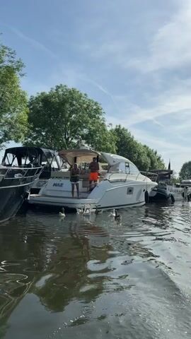 The camera, likely on a boat, faces a river lined with parked boats along the bank on a sunny day with a few clouds in the sky. The water reflects the sunlight. On the stern of a white "PRESTIGE 32" boat, two dark-skinned men are present; one is standing and wearing orange shorts, while the other is sitting. A group of ducklings are near the stern of the boat, swimming in the water. The scene shifts as the camera moves further down the river. The opposite bank of the river is visible in the distance.