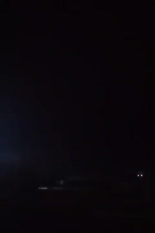 The video captures a dark, nighttime scene. Two faint white lights are visible in the distance, likely from a vehicle or distant structures. A faint blueish-white light emanates from the left side of the frame, possibly from a light source or atmospheric phenomenon. The rest of the scene is obscured by darkness, making it difficult to discern further details of the environment or any objects present.