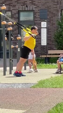The video captures a man exercising outdoors using yellow TRX suspension trainers. He is wearing a bright yellow t-shirt and black shorts. He is positioned in a plank-like pose, holding onto the handles of the trainers with his arms extended. His body is angled, demonstrating a core workout. In the background, two young girls are present. One girl, wearing a light-colored top and white shorts, appears to be standing or bending down near the exercise equipment. The other girl, in a blue top and shorts, is sitting on a wooden bench, holding a cup. A soccer ball is visible on the ground near the bench.

The setting appears to be a modern outdoor fitness area, likely part of a residential complex, given the brick building and well-maintained landscaping. The ground is paved with a textured surface, and there are patches of green grass. The sun is shining brightly, suggesting it is a clear day, likely daytime. The man's exertion is evident, but no strong emotions are discernible. The overall atmosphere seems to be one of casual recreation and fitness.

Behind the man, a sign with a pictogram of a person on a swing or slide is mounted on a pole. The building in the background has windows and a door, and a tree is visible to its right. The presence of children and the outdoor gym equipment indicate a community space designed for various activities. There is no visible text that is clear enough to read.