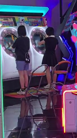 Two individuals, viewed from behind and slightly to the side, stand on separate dance game machines. The machine on the left is white with a large circular screen and bright blue overhead lighting. The individual on the left wears a black t-shirt and light blue shorts. The machine on the right is also white with a large circular screen. The individual on the right wears a black dress with a white collar. Both individuals appear to be interacting with the games, with their hands raised and possibly touching the screens. Two orange chairs with metallic frames are positioned in front of the machines, their reflections visible on the highly polished floor. The environment is dimly lit, with the bright lights of the arcade machines creating a vibrant atmosphere. Neon pink and blue lights are visible in the background, suggesting a typical arcade setting. The floor is reflective, mirroring the machines, chairs, and individuals.