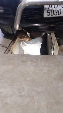 A person is working under a car in a garage. The car is a dark color and has a license plate that reads "BY AO 5030". The person is wearing a white shirt and is partially visible in the frame. The garage appears to be a repair shop, with concrete floors and metal support beams. The person is using tools to work on the car.