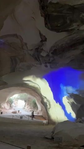 A person walks up a set of stairs inside a large cave. The cave walls are a light tan color with dark gray and black veins. Blue and yellow light is projected onto the cave walls, creating abstract patterns. A projector is visible on the ground to the right. Outside the cave entrance, trees and vegetation are visible.
