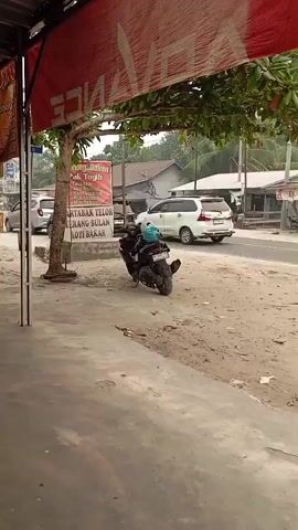 The video shows a street scene. In the foreground, there is a sidewalk or paved area. To the left, a red banner with the word "ADVANCE" is visible, hanging over a canopy. A tree with green leaves grows from the sidewalk area. Behind the tree, a sign with Indonesian text is present. A black scooter is parked on the dirt area next to the sidewalk. In the background, a road is visible with cars and motorcycles. Buildings line the other side of the road. Further back, more trees and vegetation are present, with some houses. The video then pans to the right, showing more parked motorcycles under a covered area or building. Several people are standing or walking around this area, and a woman in an orange outfit is visible. The scene appears to be an outdoor market or a street in a town.
