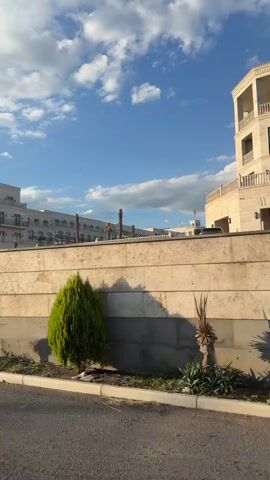 The video opens with a shot of a building with a sky full of clouds. A car drives past as the camera pans to the right, revealing more of the building's facade. The camera continues to move, showing a crosswalk and more buildings in the background. A car is parked to the right of the crosswalk. The video then shows a closer view of a bus stop shelter with reflections of the buildings in the glass. A car drives by the bus stop.