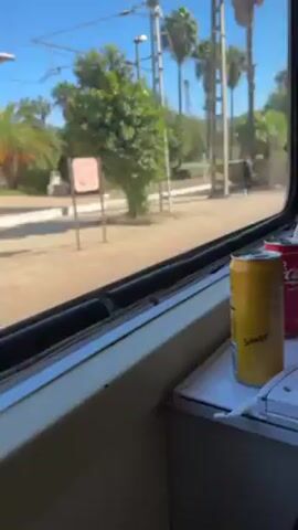 The video shows the view from a train window at a train station. The train is stationary, and the camera pans slightly to the right, showing the station platform, which is paved with a gray-brown tiled surface. Several wooden benches are visible under a covered area with a white roof and red supporting pillars. A small building, painted in off-white with gray trim, stands at the back of the platform. A sign with the name "Al-Wafa" is visible on the building. The sky is clear and blue. In the lower right of the frame, inside the train, a yellow can of Schweppes is sitting on a ledge near the window. Electrical wires run parallel to the train tracks.