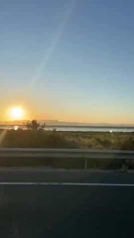 A roadside view captures a vibrant sunset over a marshy landscape and distant mountains. The camera appears to be inside a moving vehicle, with the side mirror occasionally visible, reflecting the passing scene. The sun casts a bright glare on the water and illuminates the vegetation along the roadside. The sky is clear, transitioning from a light blue to a warmer orange hue near the horizon.