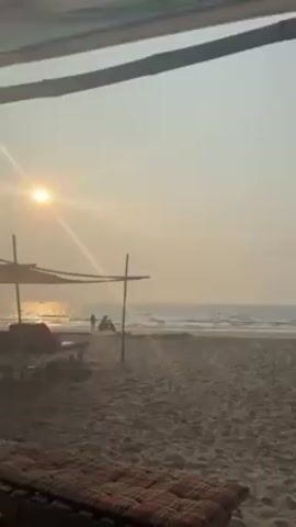 The video shows a beach at sunset. The sun is shining brightly, with its light reflecting off the water. There are a few people on the beach, one is standing and another appears to be seated on a small vehicle of some kind. There are two lounge chairs under a shaded structure on the left side of the frame, with a red and black plaid cushion on the nearest one. The shot is taken from a covered area, providing a framed view of the beach. The sky is a hazy orange color.