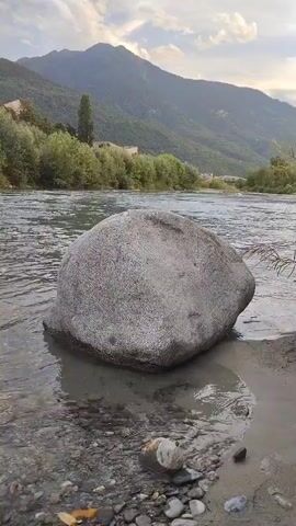 A large, gray, rounded rock sits partially submerged in the shallow, clear water of a river. The rock is positioned in the foreground, close to the camera. The river flows from left to right, its surface rippled. The background shows a lush, green, tree-lined riverbank with a few buildings visible in the distance. Beyond the trees, a mountain range rises, its peaks partially obscured by clouds. The sky above is partly cloudy. The overall scene is a tranquil, natural landscape.