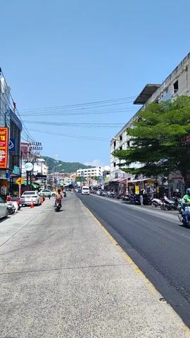 The video shows a street in a city on a sunny day. The street is lined with buildings, trees, and parked vehicles, mostly scooters. A person rides a moped down the street in the direction of the camera. A white van drives in the same direction. There are electrical wires overhead.  In the distance, there are hills or mountains.
