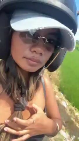 A woman wearing a black tank top, a cap, sunglasses, and a helmet is riding a motor scooter, taking a selfie video of herself as she rides, smiling at the camera occasionally. A man, wearing a helmet and shorts, is driving. They are riding on a narrow road bordered on the right by bright green rice paddies under a clear blue sky. In the distance, buildings are visible. The camera angle is from the perspective of the passenger, and the video has some shake, likely due to the motion of the scooter.