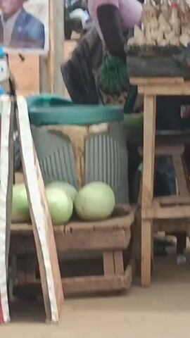 The video shows an outdoor marketplace scene. At the beginning of the video, three green watermelons rest on a wooden surface beside a beverage container and a wooden stool. As the camera pans up, a person with dark hair and a white shirt stands behind a makeshift table. The table is constructed with tires. Canned goods are stacked on the tires. A white box with the words "Cape Crisp" is also on the table. A political banner is in the background with two men's pictures and text, "ALIA/GWANDE" "THE WINNING TEAM 2027". A man in a white shirt walks in front of the table. A yellow auto-rickshaw is partially visible behind the table.
