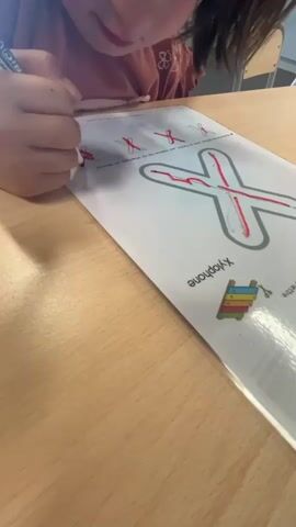 A child is drawing on a piece of paper that has the letter X printed on it. The child is holding a red marker and is tracing over the lines of the letter X. The paper also has smaller drawings of the letter X on it, as well as the word "Xylophone" and a drawing of a xylophone. The child is sitting at a wooden table, and the background is blurry but appears to be a classroom or office setting.