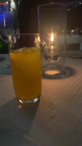 The video opens with a close-up of a glass of orange juice next to a lit candle. The shot is slightly out of focus. The camera quickly pans towards an outdoor nighttime view featuring a body of water illuminated with purple lights in the distance. The view is partially obscured by a glass railing and some shrubbery in the foreground. The scene appears to be from an elevated outdoor dining area.