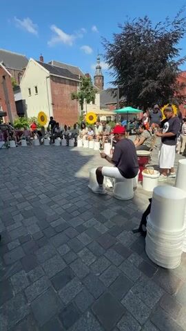 The video captures an outdoor drum circle performance on a cobblestone plaza. A group of people, including children and adults, are seated in a circle, each with a white bucket in front of them, which they are using as drums. Most participants are holding drumsticks and actively drumming. In the foreground, a young girl in a pink dress is playing a drum with two sticks. A stroller is next to her. A man in a red hat and white shorts is seated on a bucket to the right, also drumming. The background features brick buildings, a church steeple, and trees, indicating an urban setting under a clear blue sky with scattered clouds. The overall atmosphere appears to be a community event or performance.