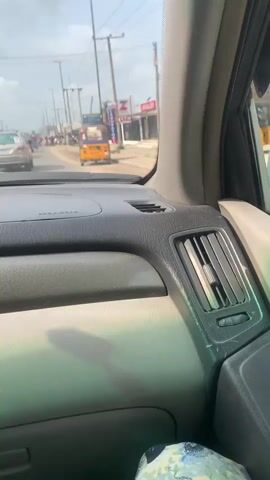 The video, shot from inside a vehicle, captures a street scene on a cloudy day. The dashboard of the vehicle is visible in the foreground, with an air vent on the right side. Ahead, a road extends into the distance, with several vehicles, including cars and a yellow tricycle, in traffic. Buildings line both sides of the road, and power lines and streetlights are visible overhead.