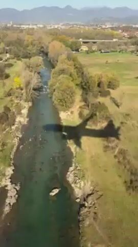 A drone camera follows the shadow of an airplane as it approaches a runway. The terrain below changes from a river, to grass, to a road with cars, to the runway itself. In the background is a city skyline and mountains.