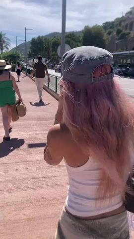 A person with pink hair, wearing a white tank top and a patterned skirt, walks away from the camera down a paved walkway. They are wearing a grey baseball cap and carrying a black purse. To their left, another person in a green dress and a straw hat walks ahead, carrying a straw bag. Further ahead, a person in a brown shirt and light-colored pants walks away from the camera. The walkway is bordered by a green fence on the right and a sidewalk with palm trees and other vegetation on the left. Cars are parked to the right of the fence, and a hillside covered in trees is visible in the background. The sky is blue and clear, indicating a sunny day.