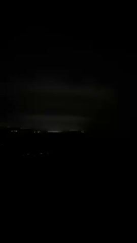 The video shows a distant cityscape at night. The horizon line is dimly lit with city lights. The sky is dark and cloudy. The video is somewhat grainy. There are indiscernible voices in the background.