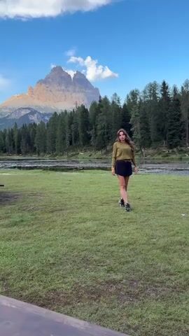 A woman with long brown hair, wearing a long-sleeved olive green top and a black skirt, stands in a grassy field. She is looking towards the camera and then turns her head to the right while touching her hair. Behind her is a body of water, a line of pine trees, and a large mountain range with rocky peaks under a partly cloudy blue sky. The foreground shows a blurry wooden object.