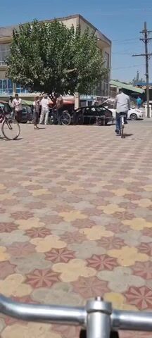 The video shows a street scene from the perspective of someone riding a bicycle. In the background, there are several people standing near some parked bicycles and a car with its trunk open. A man is riding a bicycle in the distance, away from the camera. The ground is paved with colorful tiles. The overall atmosphere appears to be a sunny day in a public area.