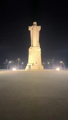 A tall, stone statue of a robed figure stands illuminated at night in a wide, open plaza. The statue's base is a large, multi-tiered structure, and the figure itself is depicted with arms outstretched. The plaza is paved and appears empty, with palm trees visible in the background. Several lights on poles line the plaza, casting a warm glow. The sky is dark, indicating nighttime. The overall scene is one of solemnity and grandeur.